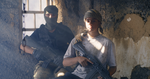 Young ethnic men with rifles looking at camera while sitting near window inside grungy building during war