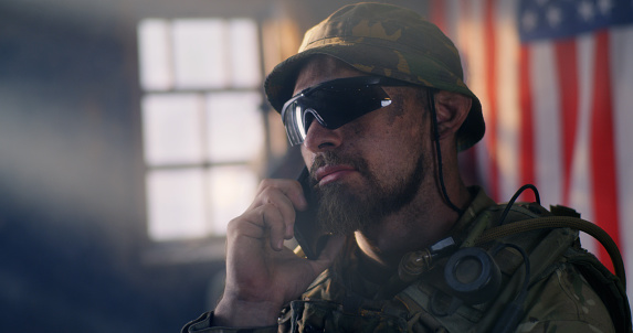 Bearded man in camouflage and sunglasses smiling and speaking on smartphone against USA flag during war