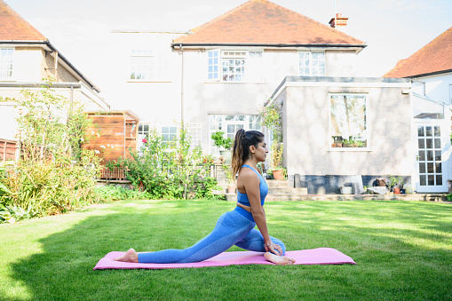 Side view of woman in late 20s wearing leggings and sports bra doing Pada Rajakapotasana yoga pose on exercise mat in backyard.