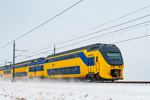 Blue and yellow Dutch intercity train of the Nederlandse Spoorwegen (NS) driving through the snow on a cold winter day. Snow is blowing up from the ground as the train is passing.