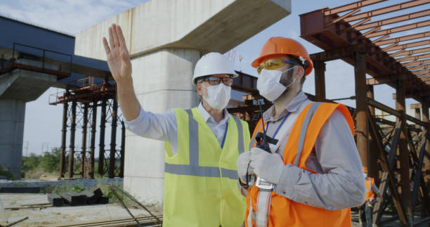 Contractors in masks talking during work Team of builders in uniform and medical masks speaking with each other and giving orders on radio while working on construction site during epidemic medium shot stock pictures, royalty-free photos & images