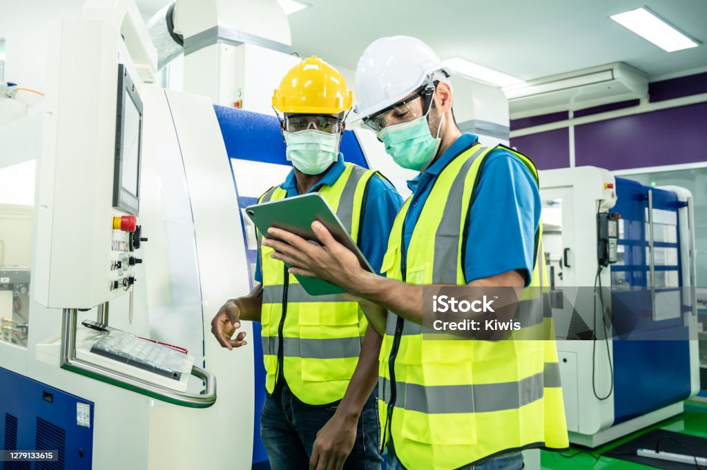 Group of interracial worker people wearing protective face mask in production factory due to covid pandemic crisis. Engineers care of health working in industry looking and operating the machine. Safety Stock Photo