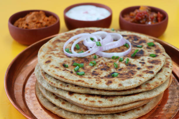 Stock photo showing a close-up view of aloo paratha pile on a metal, dimpled tray with dishes of tasty chutney, pickle dip and raita (yoghurt based sauce), terracotta pots. Savoury flatbreads with garam masala potato filling, a popular breakfast in India. imagen de bandeja de metal con pila de aloo paratha cubierta con aros de cebolla roja y espolvoreado de cebollinos picados, pan plano salado relleno con puré de garam masala papas, cuencos de chutney, chapuzón de pepinillo y raita (yogur), fondo amarillo
