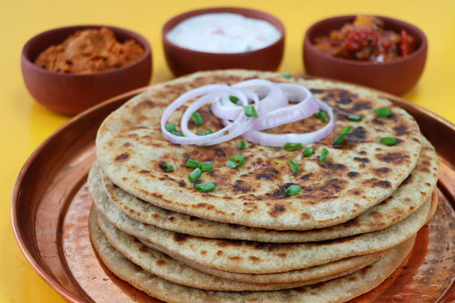 Stock photo showing a close-up view of aloo paratha pile on a metal, dimpled tray with dishes of tasty chutney, pickle dip and raita (yoghurt based sauce), terracotta pots. Savoury flatbreads with garam masala potato filling, a popular breakfast in India.