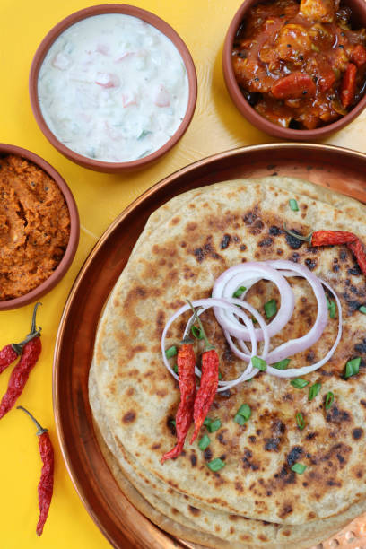 Image of metal tray with with aloo paratha pile topped with chillies, red onion rings and sprinkle of chopped chives, savoury flatbread stuffed with mashed garam masala potatoes, chutney, pickle dip and raita (yoghurt), elevated view stock photo