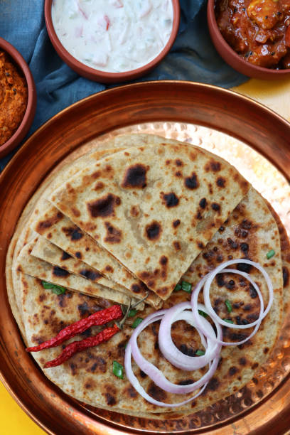 Stock photo showing an elevated view of triangular slices of aloo paratha pile on a metal, dimpled tray beside dishes of tasty chutney, pickle dip and raita (yoghurt based sauce) and a tea towel. Savoury flatbreads with garam masala potato filling, a popular breakfast in India. imagen de bandeja de metal con rodajas triangulares de pila de aloo paratha cubierta con chiles, aros de cebolla roja y espolvorear cebollino picado, pan plano salado relleno de puré de garam masala papas, chutney, pickle dip y raita (yogur), vista elevad