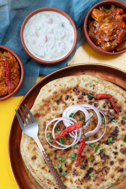 Stock photo showing an elevated view of aloo paratha pile on a metal, dimpled tray beside dishes of tasty chutney, pickle dip and raita (yoghurt based sauce), metal fork and a tea towel. Savoury flatbreads with garam masala potato filling, a popular breakfast in India. imagen de bandeja de metal con pila de aloo paratha, chiles, aros de cebolla roja y espolvorear cebollino picado, pan plano salado relleno con puré de garam masala papas, chutney, pickle dip y raita (yogur), tenedor de metal, fondo amarillo, vista elevada