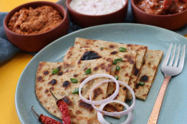 istock Image of green plate, triangular slices aloo paratha, chillies, red onion rings and sprinkle of chopped chives, savoury flatbread stuffed with mashed garam masala potatoes, chutney, pickle dip, raita (yoghurt), metal fork, focus on foreground 1279135175