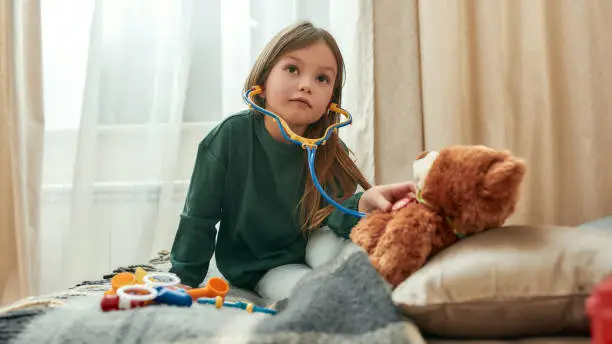 A little cute girl listening to a heartbeat of a teddybear through a toy stethoscope sitting on a sofa in a bright guestroom at home alone. Childrens leisure activities