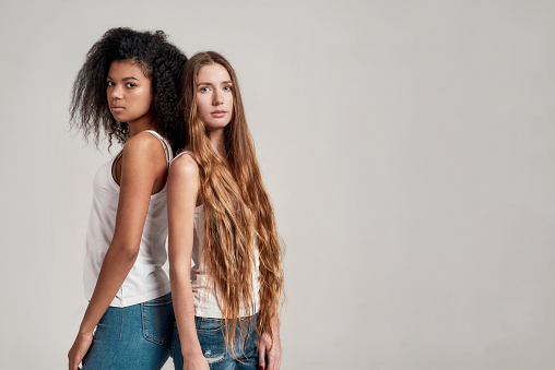 Two young diverse women wearing white shirts looking at camera while standing together isolated over grey background. Diversity, beauty concept. Horizontal shot