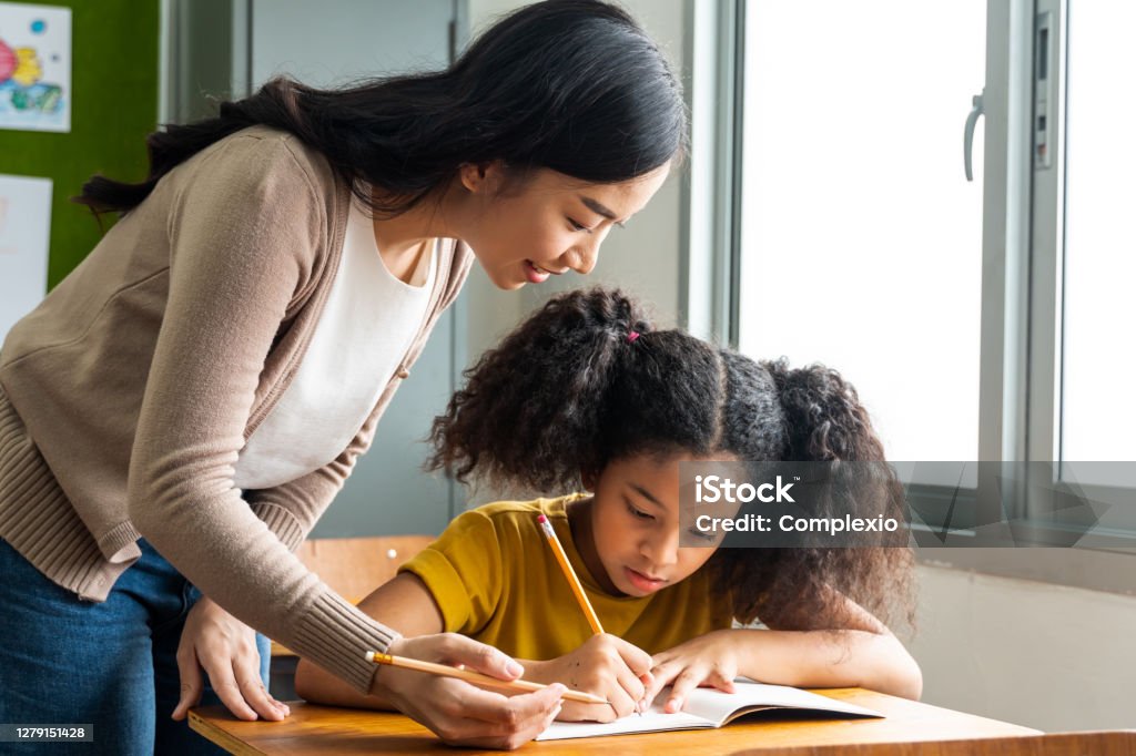 Asian school teacher assisting female student in classroom. Young woman working in school helping girl with her writing, education, support, care Asian school teacher assisting female student in classroom. Young woman working in school helping girl with her writing, education, support, care. Teacher Stock Photo