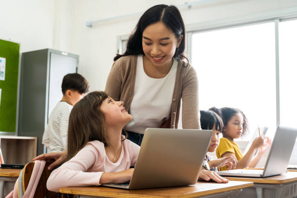 istock Young Asian teacher watching Caucasian school girl using computer in school classroom, digital native, technology, learning, guidance, education. Female instructor and student smiling 1279151455