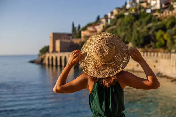 Horizontal backview portrait of a fit female wearing green top and straw hat and standing in front of Alanya castle wall and medieval shipyard