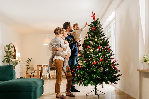 Young family of four people is standing in their living room next to the christmas tree and decorating it. They are wearing winter sweaters. Mother is holding one son and father the other. The youngest son is reaching towards christmas star on the tree. They are smiling and having a good time. Bright and cozy living room. Horizontal photo with copy space.