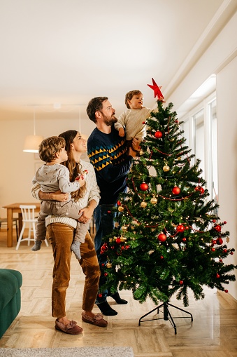 Young family of four people is standing in their living room next to the christmas tree and decorating it. They are wearing winter sweaters. Mother is holding one son and father the other. The youngest son is putting the christmas star on the tree. They are smiling and having a good time. Bright and cozy living room. Vertical photo with copy space.
