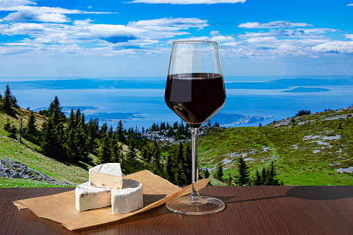 Glass of red wine with cheese against mountains background. View of blue sea and green valley in the mountains.