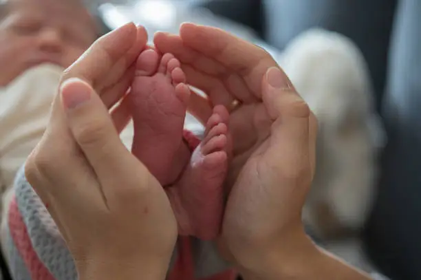 Feet of a new born baby in the middle of her mothers hands protecting the new life in the blurred background you can see the baby sleeping