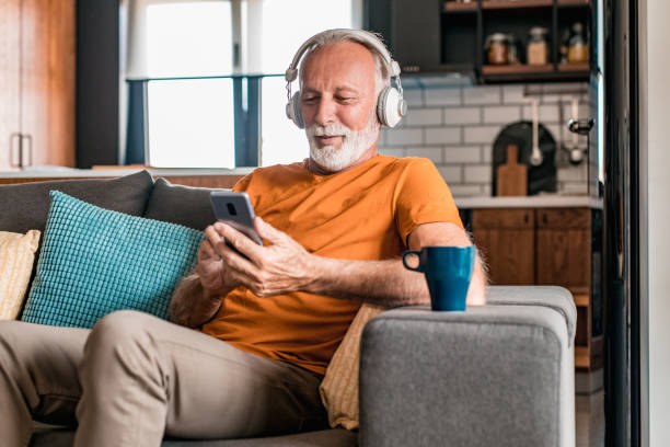 homme aîné avec des écouteurs à la maison utilisant le téléphone intelligent - only senior men audio photos et images de collection