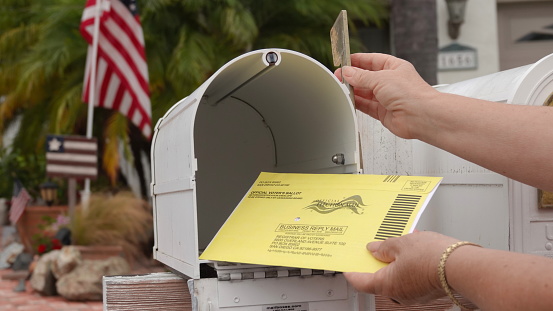 Close up of an American mailbox with flags in the background, older woman's hands returning mail-in election ballot. Illustrative editorial taken in Vista, CA / USA on October 8, 2020.