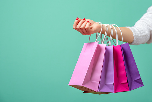 Colored paper bags on a woman's hands against a blue background. Young woman holding on her hand pink and purple shopping bags.