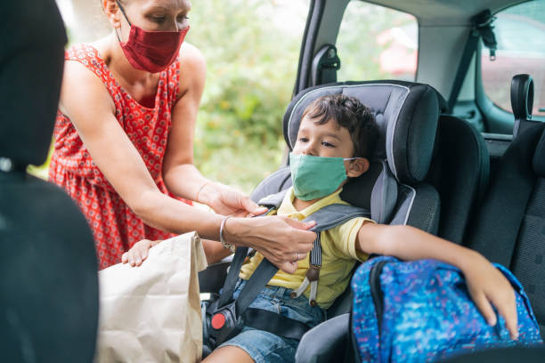 istock Boy wearing protective face mask sitting in the car for his first day back to school while being secured in his car seat by his mother, during COVID-19 1279212021