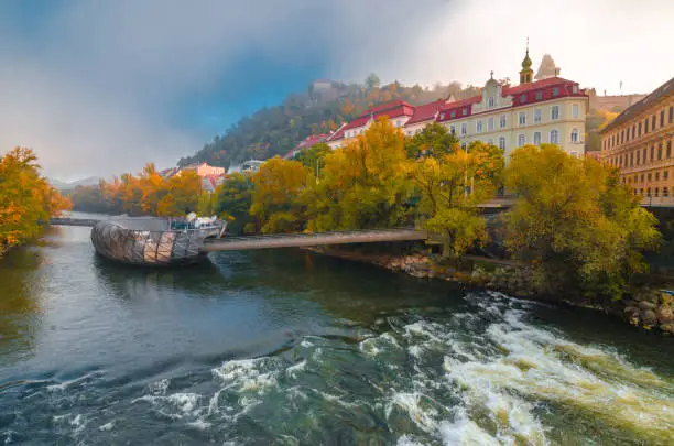 Mur river in autumn, with Murinsel bridge and old buildings in the city center of Graz, Styria region, Austria.