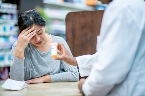 A depressed, overwhelmed woman sits at the pharmacy counter. There is a pill bottle labelled \