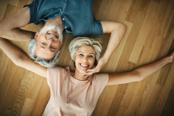 Loving mid aged couple at home. Closeup top view of early 50's and 60's couple laying on the hardwood floor, smiling and looking at camera. grey hair on floor stock pictures, royalty-free photos & images