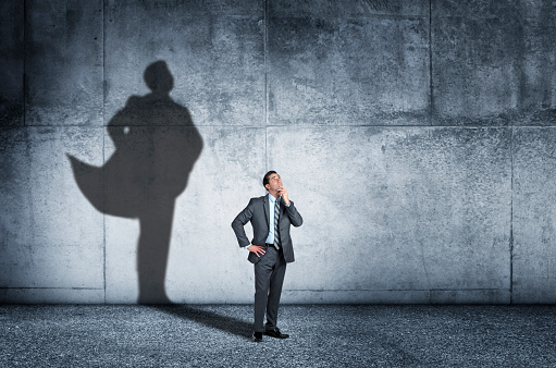 A man strikes a contemplative pose as his shadow that is cast on the wall behind him shows him in a confident stance and wearing a cape.