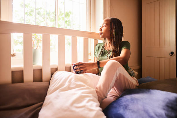 young woman looking out of her bedroom window while drinking coffee - braids african descent women pensive imagens e fotografias de stock