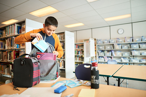 Shot of a boy standing in classroom packing his schoolbag after the class