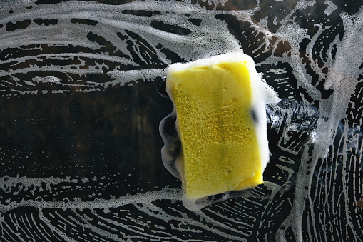 Hand in a black glove washes the glass with a foam sponge with foam on a dark background.