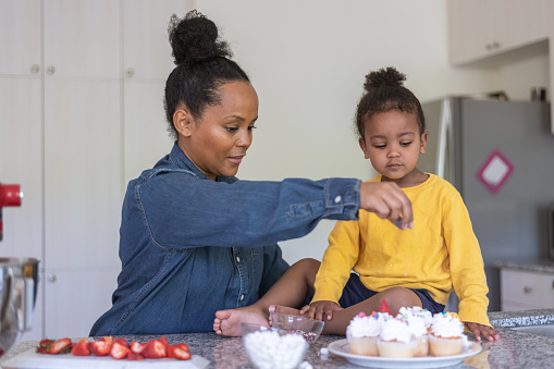 An ethnic mother makes cupcakes with her young son, who is sitting on the counter. The cupcakes are setting on a plate and look delicious!