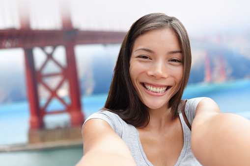 Selfie girl on San Francisco Golden bridge travel. Cute young Asian woman adult taking picture with her smartphone during summer vacation in front of the famous American attraction, California, USA.