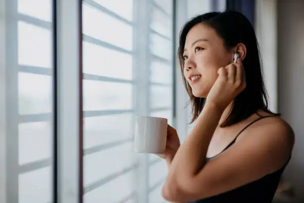 Young asian woman listening to music with wireless headphone and drinking coffee, enjoying a peaceful moment at home