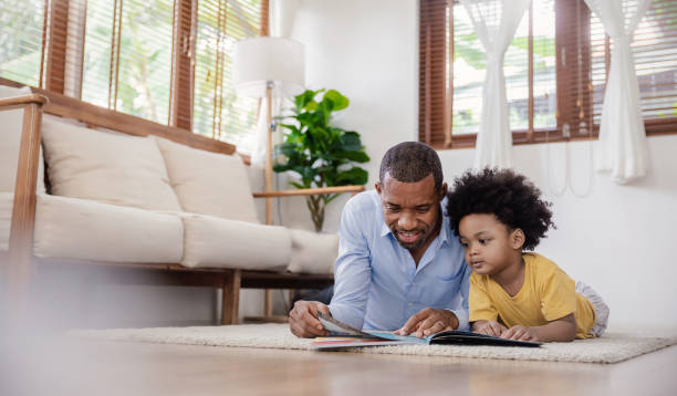 portrait of african american father hold story book reading teaching little boy in living room. home school learn from teacher, education happy family, fatherâs day concept - family reading african descent book imagens e fotografias de stock