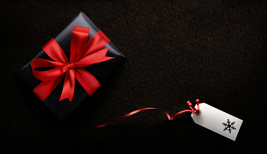 A Christmas present wrapped in black paper and red ribbon with a blank tag, card against a dark background from above.