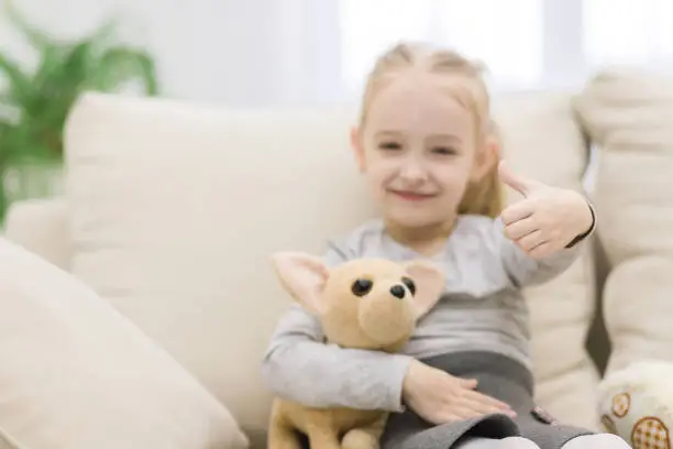 Photo of Beautiful little girl is sitting on sofa with teddy bear and shows thumb up.