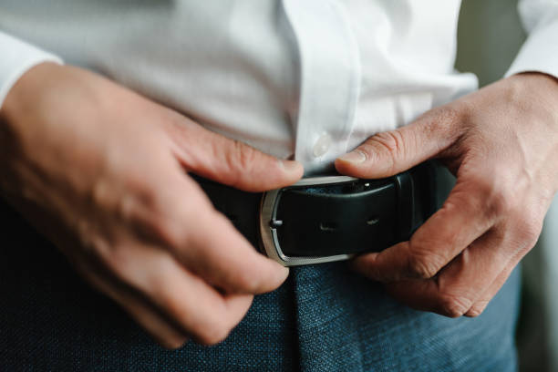 a man in pants and shirt buttoned a black leather trouser belt. groom dress up a belt with a buckle. businessman wear leather stylish belt. close up. - belt personal accessory leather fashion imagens e fotografias de stock