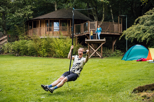 A wide shot of a young man holding an action camera on a zip line swing smiling.