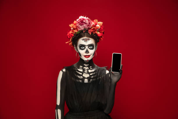 Young girl in the image of Santa Muerte, Saint death or Sugar skull with bright make-up. Portrait isolated on studio background. Holding smartphone. Young girl like Santa Muerte Saint death or Sugar skull with bright make-up. Portrait isolated on red studio background with copyspace. Celebrating Halloween or Day of the dead. muerte stock pictures, royalty-free photos & images