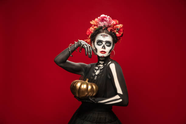 Young girl in the image of Santa Muerte, Saint death or Sugar skull with bright make-up. Portrait isolated on studio background. Pumpkin golden. Young girl like Santa Muerte Saint death or Sugar skull with bright make-up. Portrait isolated on red studio background with copyspace. Celebrating Halloween or Day of the dead. muerte stock pictures, royalty-free photos & images