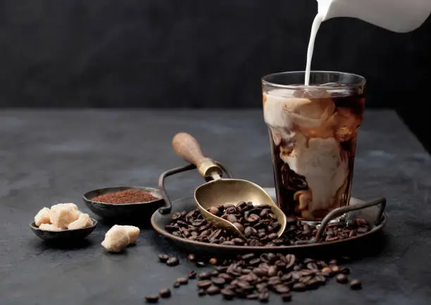 Pouring fresh milk into glass of iced black coffee on tray with beans and ground coffee with cane sugar and vintage shop on black background. Space for text