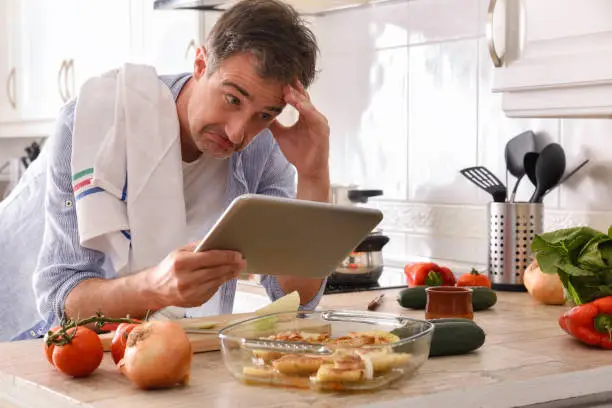 Novice chef looking at a tablet with an inept face resting his elbow on the kitchen bench touching his forehead
