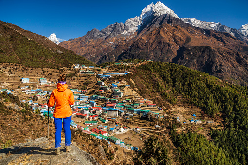 Young woman, wearing orange jacket, is standing on the top of a rock and looking at Namche Bazaar village in Mount Everest National Park. This is the highest national park in the world, with the entire park located above 3,000 m ( 9,700 ft). This park includes three peaks higher than 8,000 m, including Mt Everest. Therefore, most of the park area is very rugged and steep, with its terrain cut by deep rivers and glaciers.