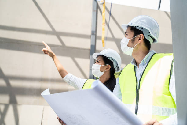 Asian workers people wearing protective face mask onsite of architecture due to covid pandemic crisis. Male and female engineers holding drawing working on construction side discussing about project. Asian workers people wearing protective face mask onsite of architecture due to covid pandemic crisis. Male and female engineers holding drawing working on construction side discussing about project. slow motion face stock pictures, royalty-free photos & images