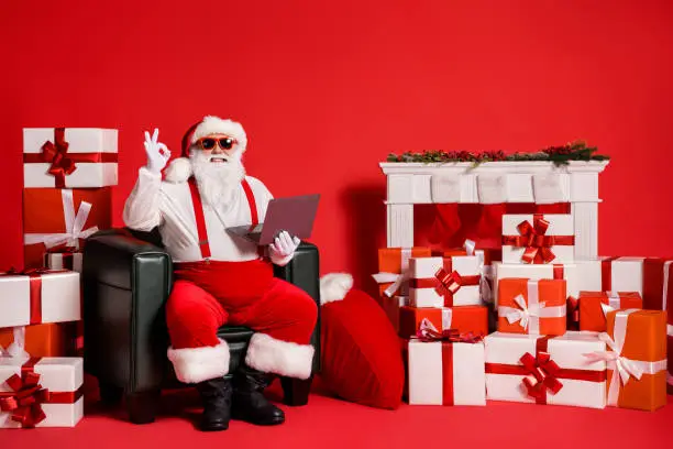 Portrait of his he nice, attractive funky fat bearded cheerful Santa using laptop sitting in armchair showing ok-sign advert agree isolated bright vivid shine vibrant red color background