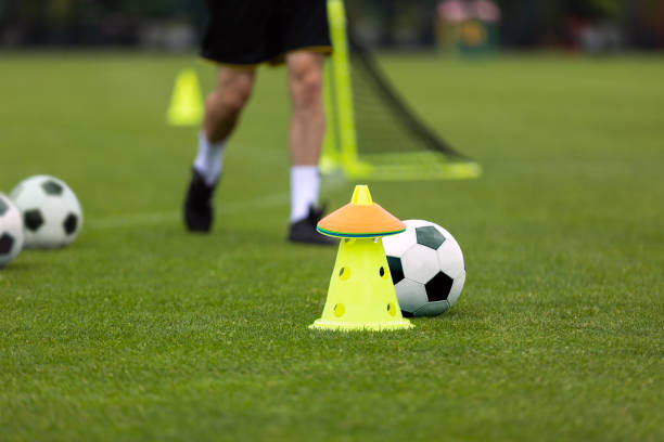 joueur adulte de football sur l’unité d’entraînement. footballeur en cours d’exécution à l’entraînement. cône jaune de football, marqueurs d’entraînement et ballon de football au premier plan. champ d’herbe brouillé à l’arrière-plan - football helmet playing field american football sport photos et images de collection