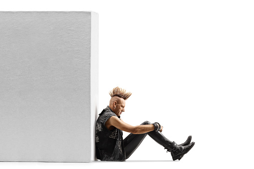 Punk rocker with a mohawk sitting on the ground and leaning on a white wall isolated on white background