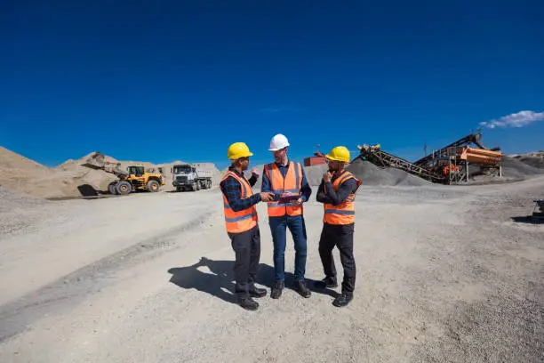 Open- pit mine engineer and workers wearing protective clothes and helmets standing in front of pile of gravel and excavator. Manager holding clipboard and discussing with team. Wide angle view.